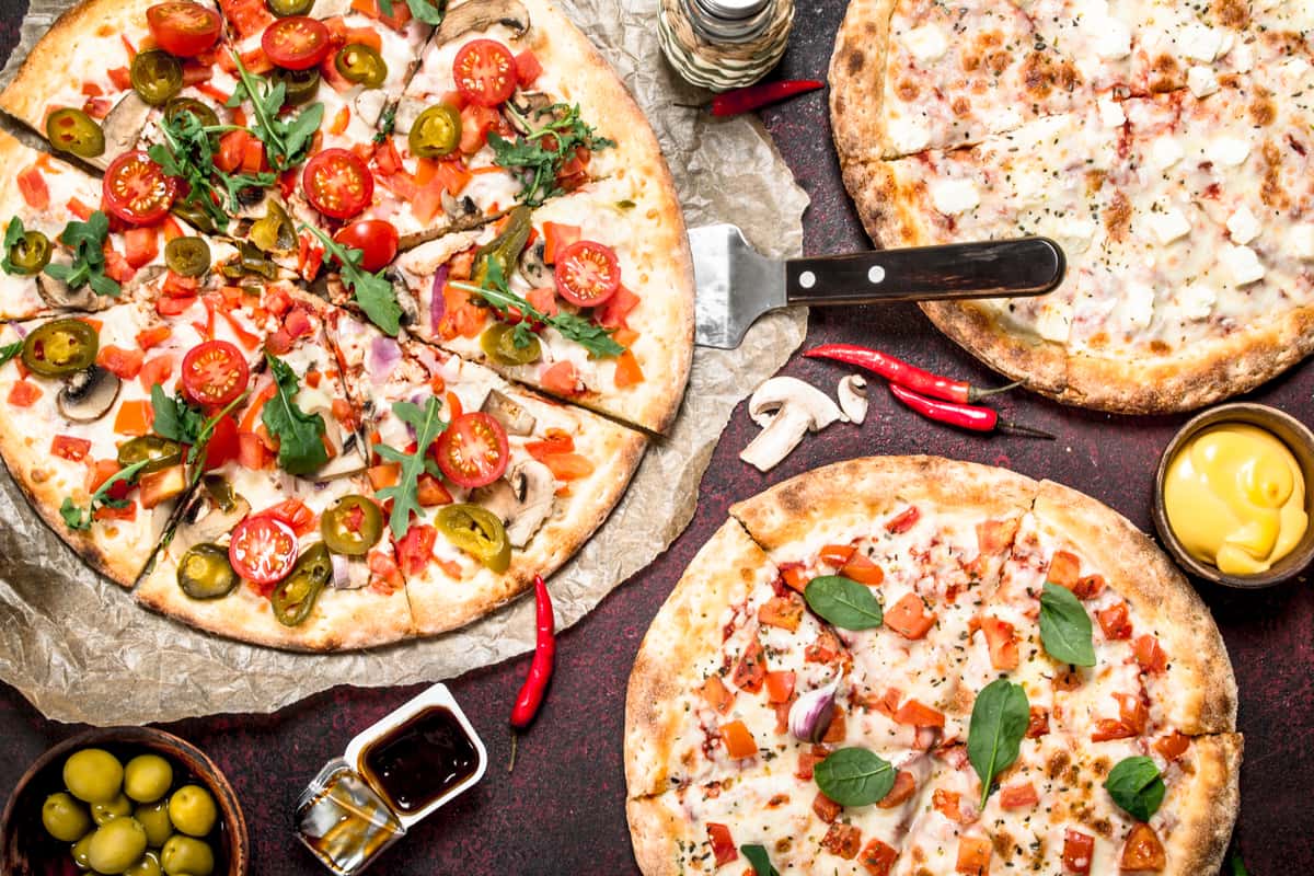 Old Shawnee Pizza's three pizzas with various toppings sit on a dark surface alongside pizza-making ingredients.