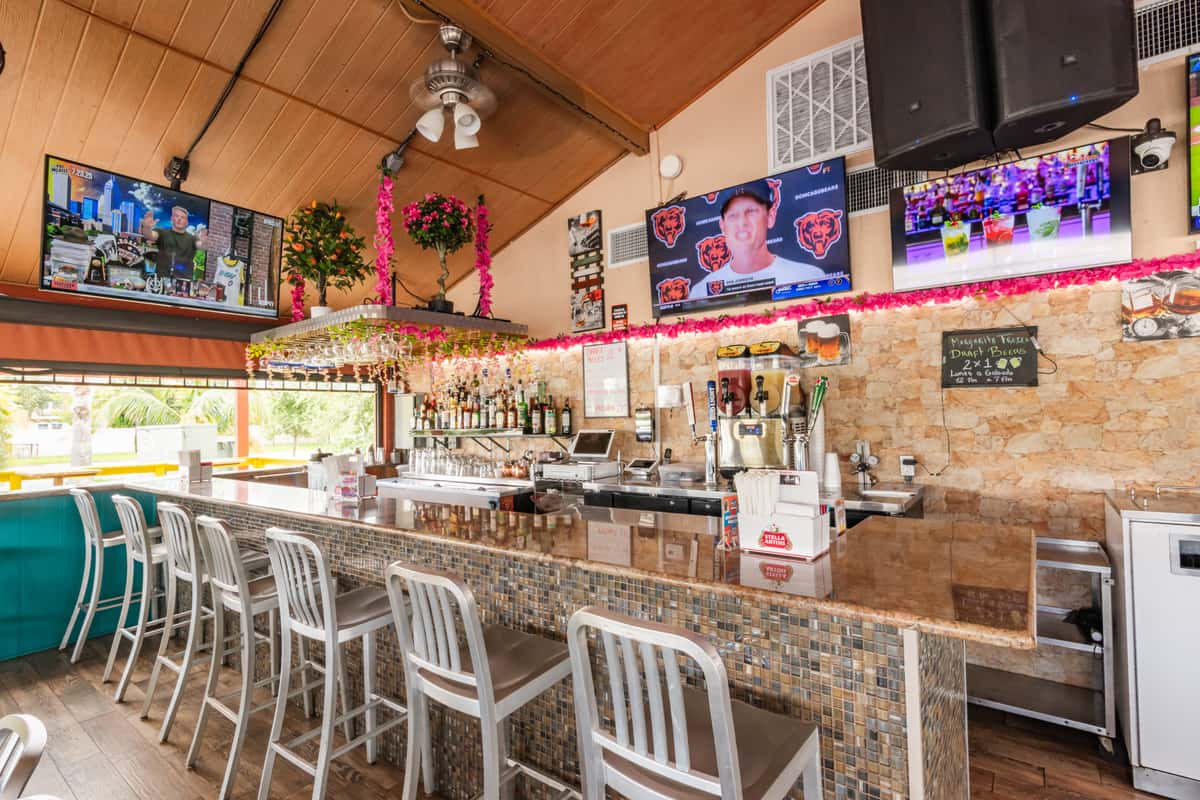Lively Caribbean bar interior with granite countertop, metal barstools, overhead TVs, and tropical floral decor—perfect setting for cocktails, sports, and casual Latin dining.