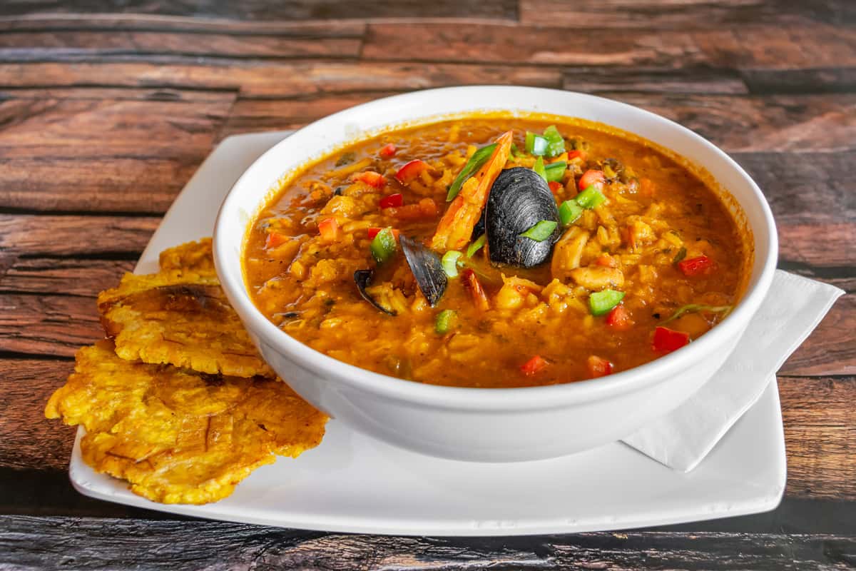 Bowl of Caribbean seafood stew loaded with shrimp, mussels, and rice in a vibrant tomato-based broth, garnished with bell peppers and scallions, served with crispy tostones—flavor-packed Dominican-style asopao de mariscos.