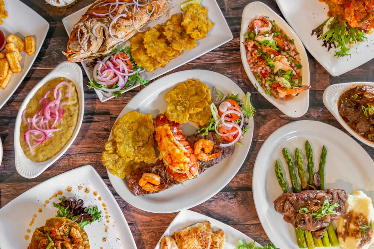 Best Dominican food near me - Colorful Dominican feast featuring mofongo topped with shrimp, fried whole red snapper with tostones, grilled steak and lobster tail with shrimp, mangú with pickled red onions, shrimp ceviche, and churrasco steak with asparagus and mashed potatoes. Perfect representation of authentic Caribbean cuisine in a vibrant, rustic setting.