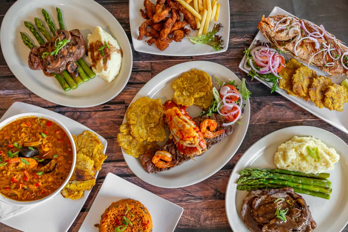 Vibrant spread of Caribbean cuisine featuring grilled lobster and steak with tostones, asopao de mariscos, fried red snapper, churrasco steak with mashed potatoes and asparagus, fried chicken with fries, and a side of yellow rice—perfect showcase of Dominican and Latin American culinary favorites.