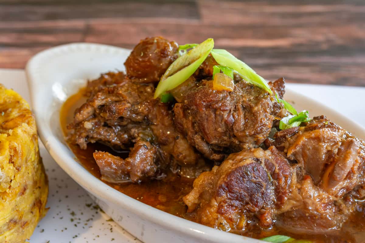 Tender braised oxtail in rich brown gravy, garnished with scallions and served with a side of mofongo—hearty and flavorful Caribbean comfort food with Dominican flair.