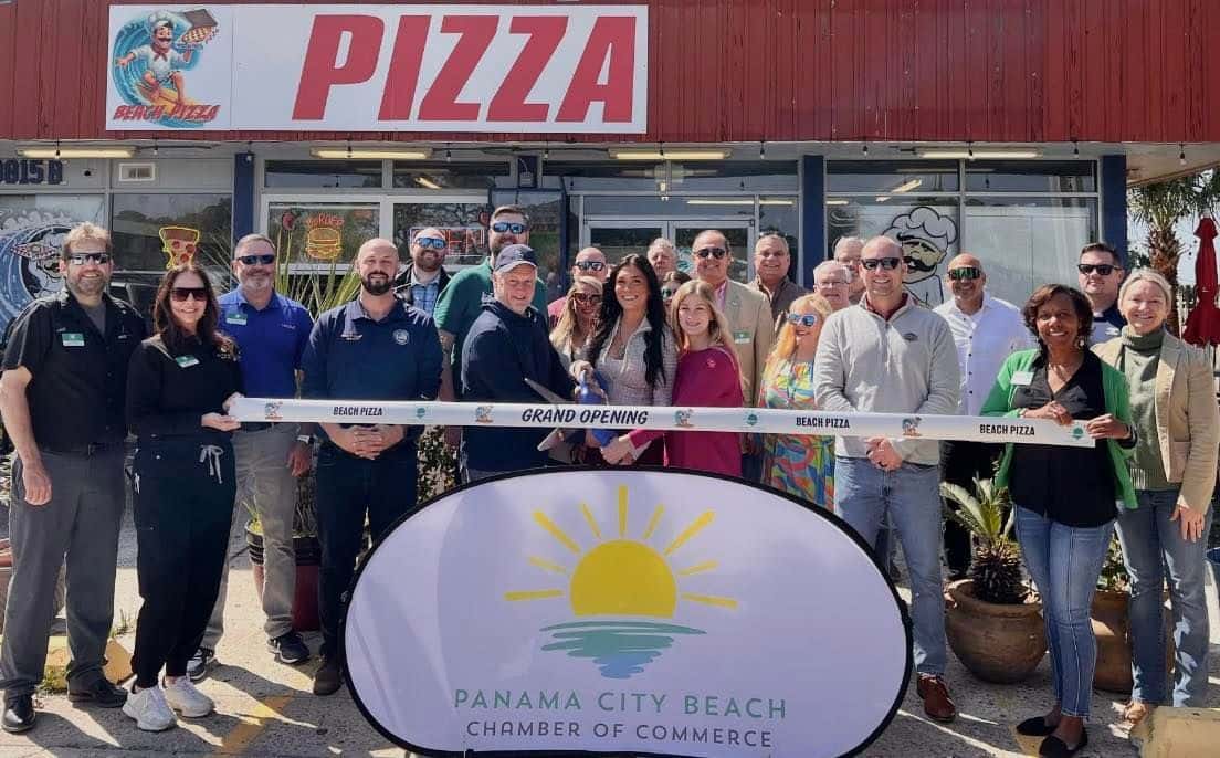 Group of people celebrating a ribbon-cutting ceremony outside Beach Pizza, with a Panama City Beach Chamber of Commerce banner.