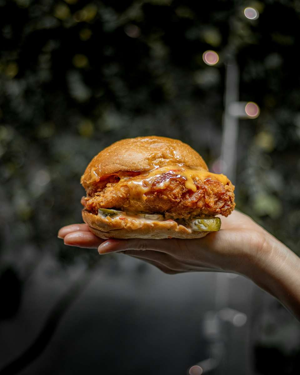 Hand holding a juicy fried chicken sandwich dripping with melted cheese and sauce, layered with pickles and lettuce on a soft bun, perfect for gourmet fast casual restaurants.