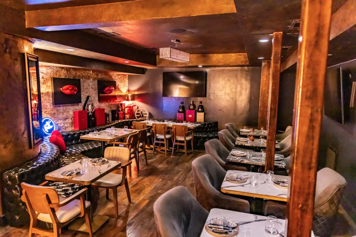 Upscale lounge-style restaurant interior with wood floors, velvet dining chairs, leather banquette seating, and softly lit tables arranged throughout the room. Warm lighting, textured walls, and modern décor create a cozy, speakeasy-inspired setting.