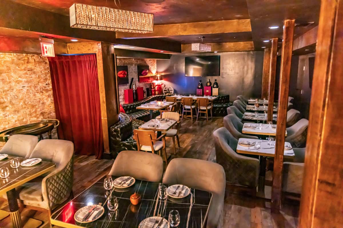 "Warm, upscale restaurant dining room with dim ambient lighting, wood-paneled walls and columns, tufted leather banquette seating, and neatly set tables. A red velvet curtain frames a doorway on the left, while a large TV hangs above a cozy lounge-style seating area in the back. The space has an intimate, speakeasy-inspired atmosphere with modern rustic décor.
