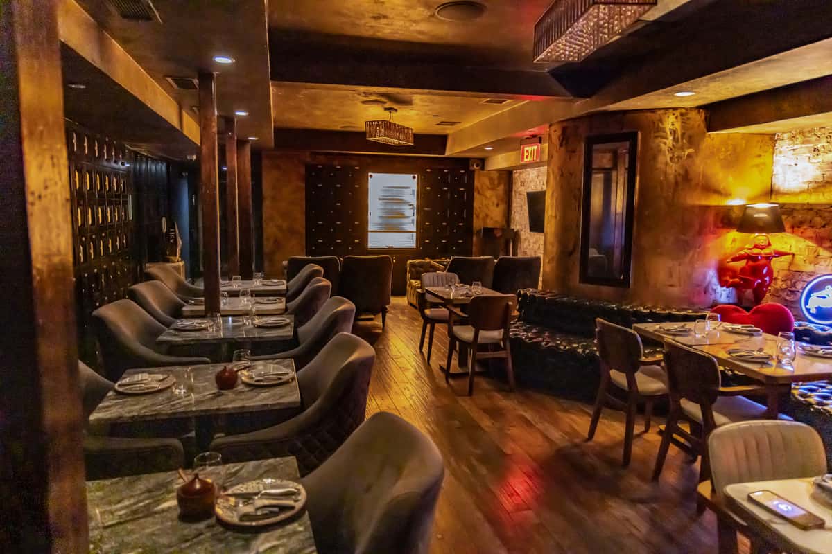 Dimly lit restaurant dining room with plush gray chairs, marble-top tables, tufted leather booths, and warm golden lighting. The space features rustic wood accents, intimate seating arrangements, and an elegant lounge atmosphere.
