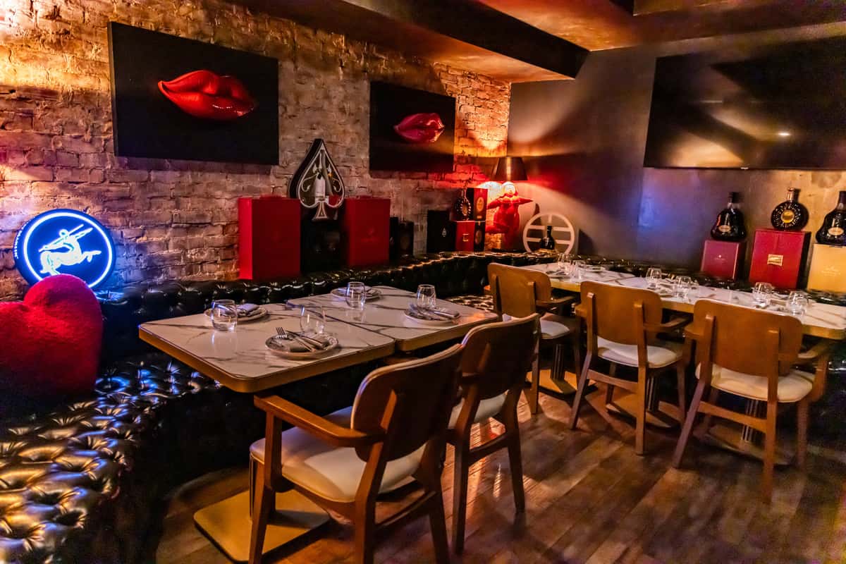 ntimate restaurant dining area with tufted black leather banquette seating, marble-top tables set with plates and glassware, and warm ambient lighting. Exposed brick walls feature bold artwork of red lips and decorative accents, creating a stylish lounge atmosphere