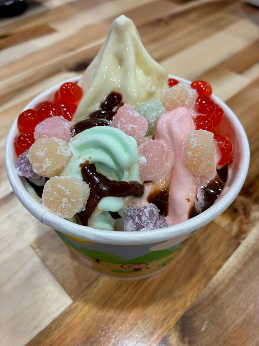 Pastel vanilla soft serve frozen yogurt topped with red popping boba, sour gummy candies, and chocolate drizzle in a polka dot cup – colorful self-serve froyo dessert perfect for a fun, family-friendly yogurt shop.