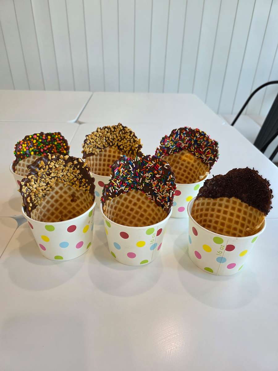 Chocolate-dipped waffle bowls coated with rainbow sprinkles, crushed nuts, and chocolate shavings, displayed in polka dot cups – gourmet ice cream shop cones ready for soft serve or frozen yogurt.