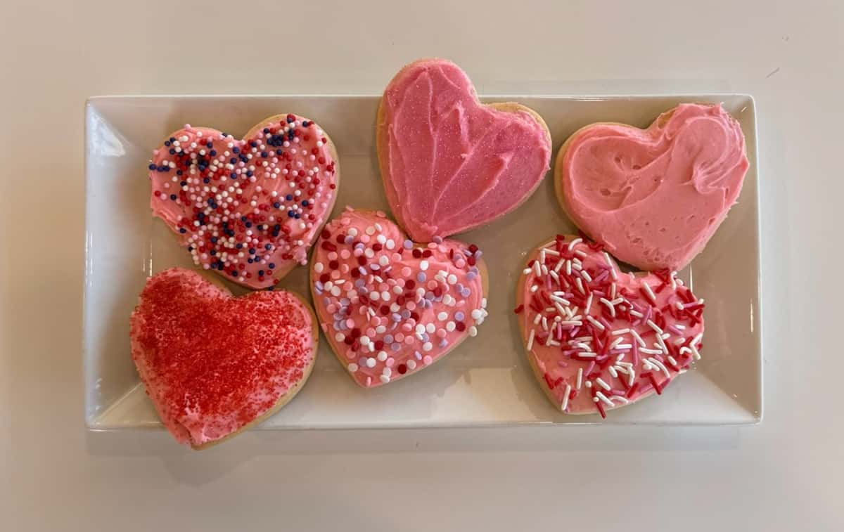 Valentine's Day heart sugar cookies decorated with pink buttercream frosting and festive red, white, and pink sprinkles, beautifully arranged on a white platter – handcrafted bakery cookies perfect for holiday dessert tables or gift boxes.