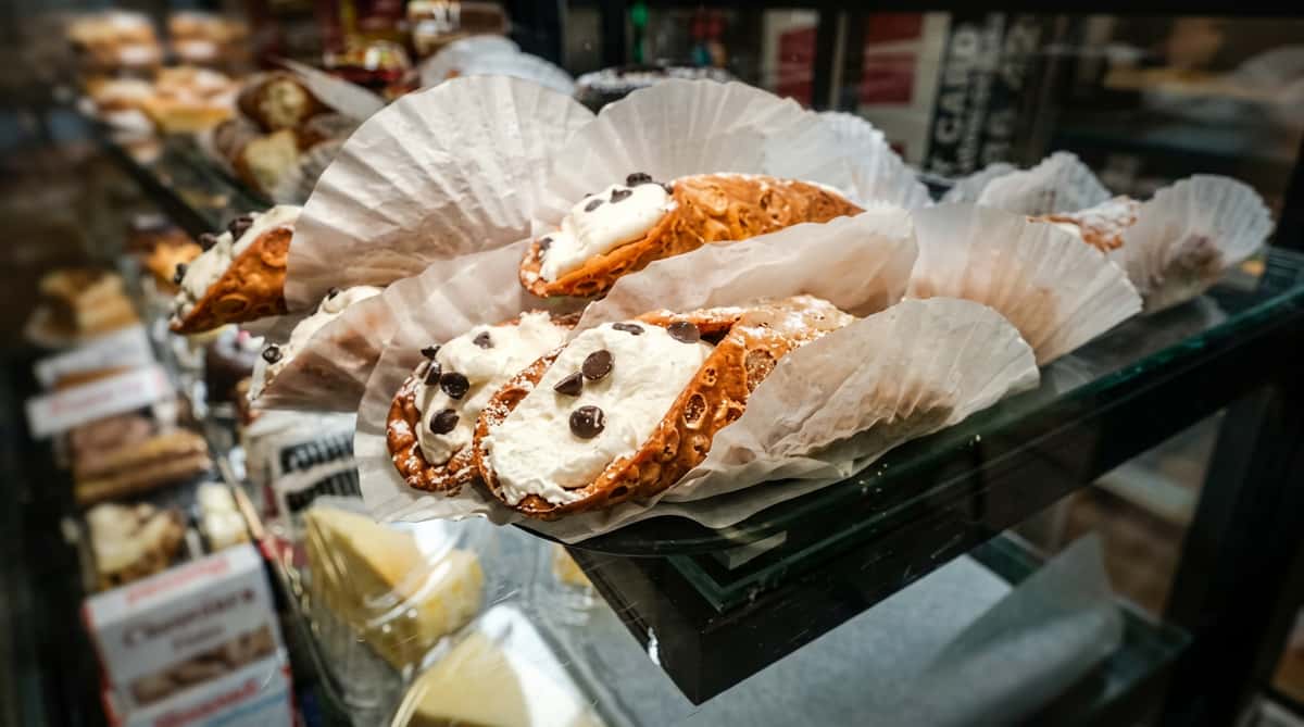 Fresh cannoli pastries filled with sweet ricotta cream and topped with chocolate chips, displayed in a bakery case with authentic Italian dessert shop ambiance.