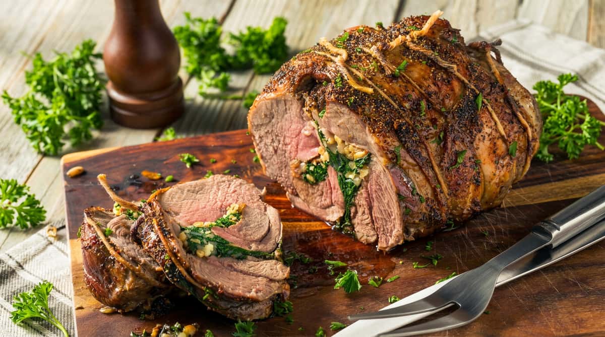 Roasted lamb leg, stuffed with visible spinach and herbs, sliced, on a wooden cutting board with a carving fork.
