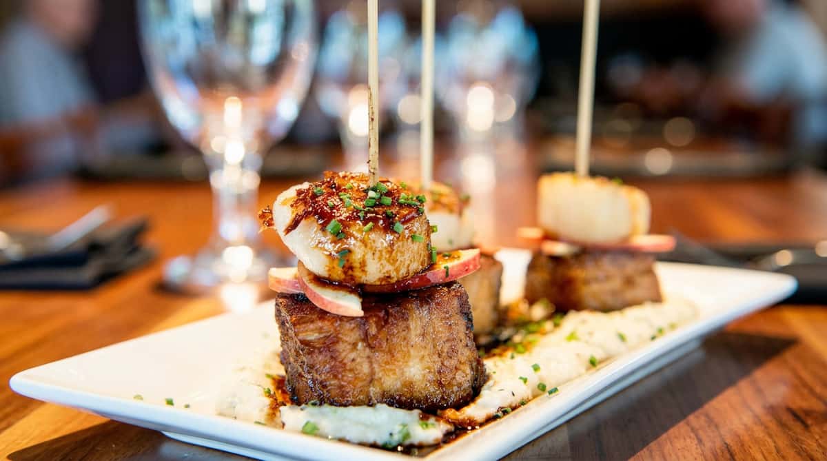 Seared scallops and pork belly skewers with apple slices, chives, and sauce on mashed potatoes.