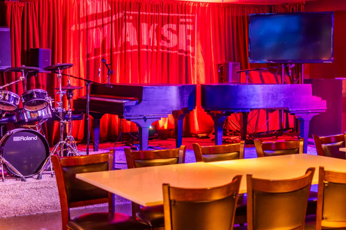 Live music stage with two grand pianos, drum kit, red curtains, and colorful lighting inside a restaurant and entertainment venue.