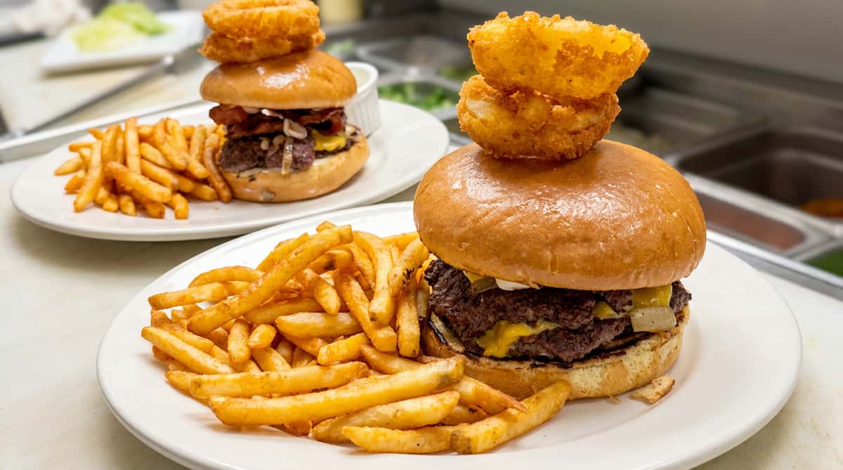 Two smash burgers with melted cheese, bacon, and grilled onions, served with golden-brown French fries and topped with onion rings.