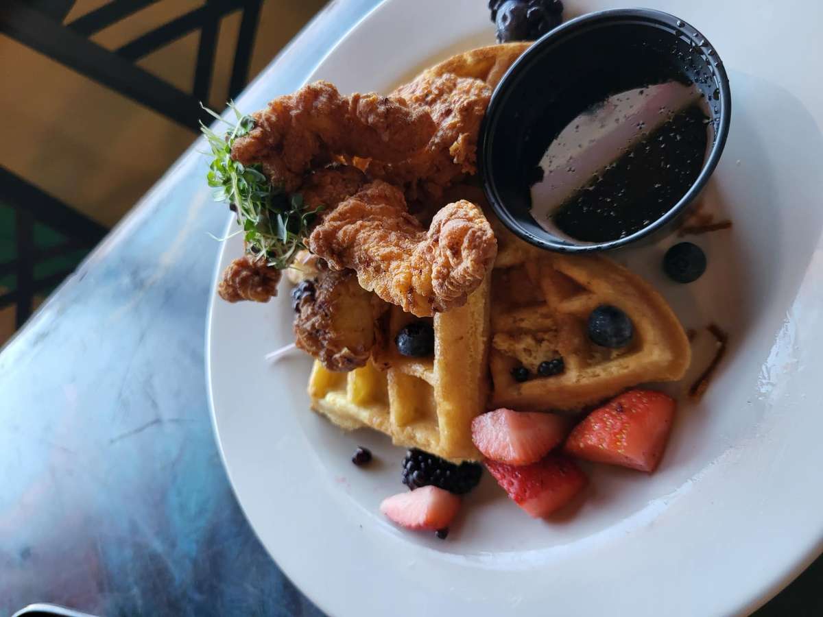 Fried chicken and waffles served with fresh berries, microgreens, and a side of syrup on a white plate.