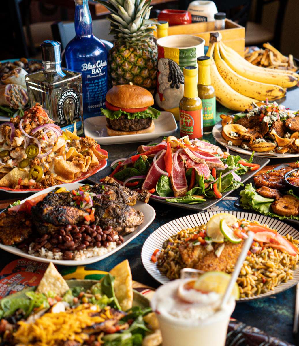 Colorful Caribbean feast with jerk chicken, seared tuna salad, nachos, tropical drinks, fried plantains, and fresh fruits.