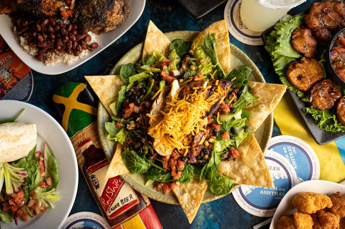 Grilled chicken taco salad served in a tortilla shell with shredded cheddar cheese, black beans, corn, lettuce, tomato, and onions. Surrounding it are Caribbean-inspired dishes including jerk chicken with rice and beans, fried plantains, and a burrito with a side salad.