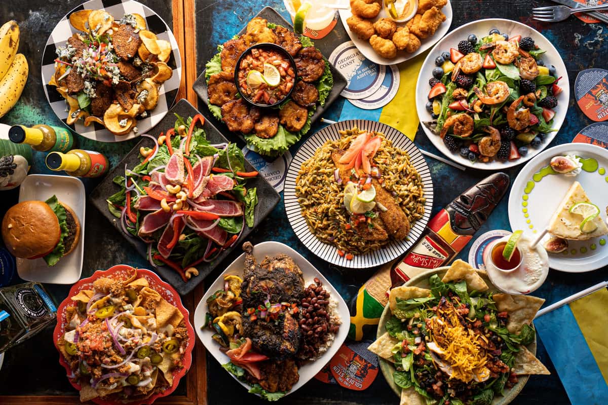 This vibrant overhead shot showcases a colorful Caribbean-inspired feast. Dishes include jerk chicken, plantains, shrimp salad, nachos, seared tuna, and a variety of other flavorful entrées and sides, all artfully arranged around a tropical, island-themed table setting. Perfectly fitting for a coastal restaurant like North Beach Bar and Grill.