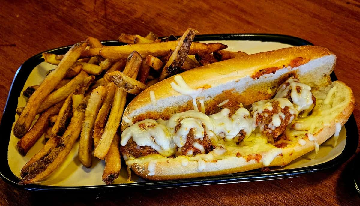 Meatball sub topped with melted cheese, served with a side of golden-brown French fries on a rectangular plate.