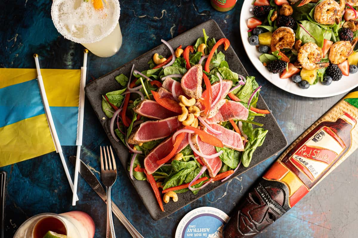 Seared ahi tuna salad with spinach, red onions, red bell peppers, and cashews, surrounded by drinks and a shrimp and berry salad.