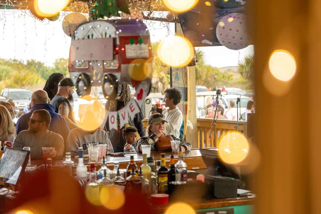 Crowd gathered at a festive beach bar decorated with lights and a "LOVE" sign, viewed through warm bokeh and a hanging camper ornament.