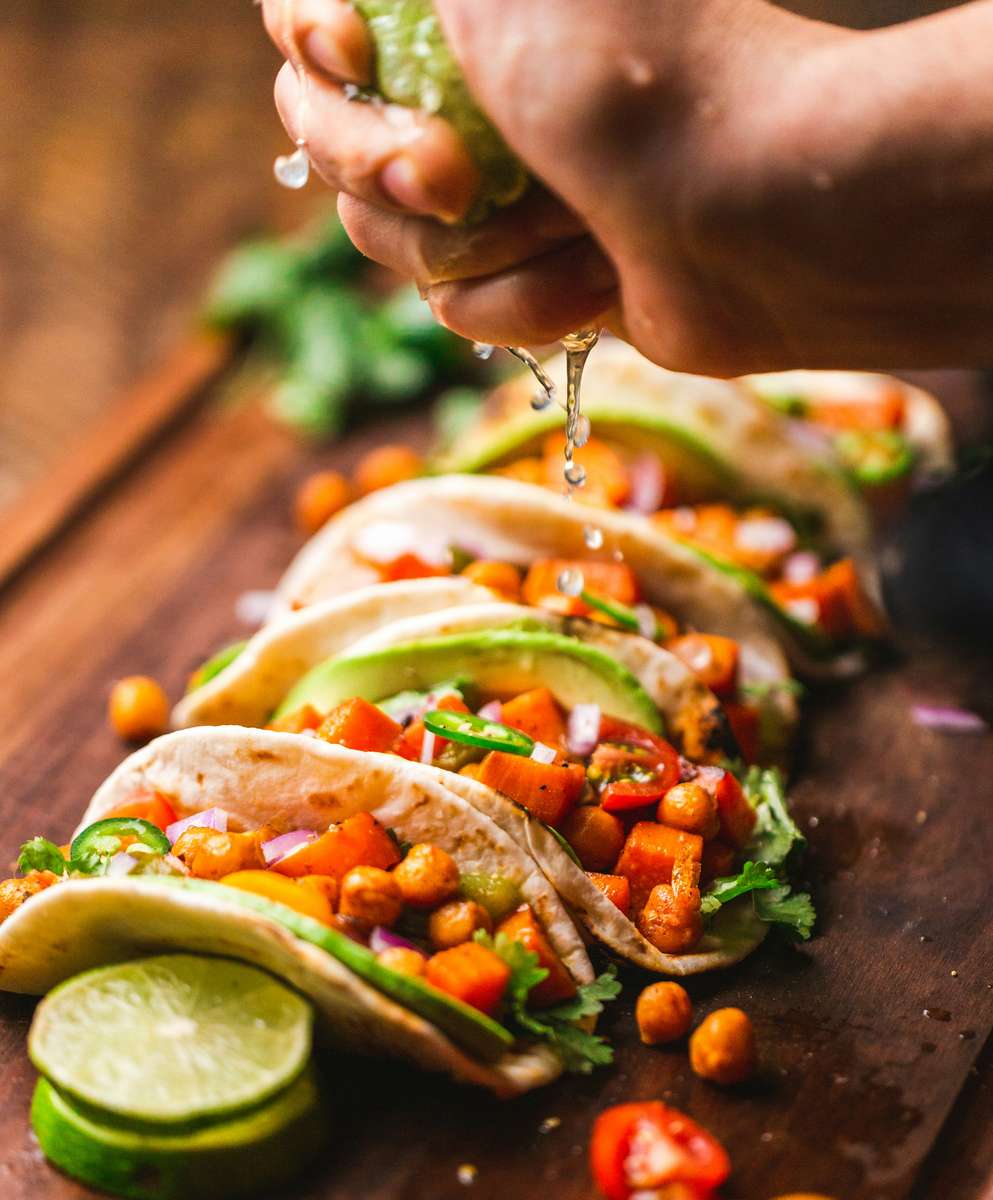 Colorful chickpea and sweet potato tacos topped with fresh avocado, jalapeños, red onions, and lime juice, served on a wooden board for a bold plant-based option.