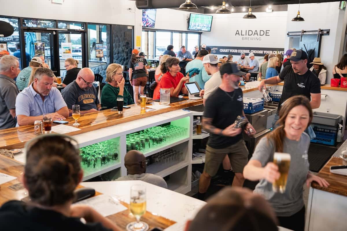 Busy Alidade Brewing taproom with guests enjoying craft beer, bartenders serving drinks, and a lively social atmosphere around the brewery bar.