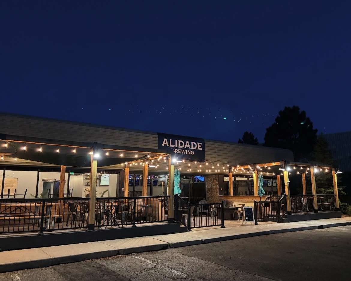 Exterior of Alidade Brewing at night with a warmly lit outdoor patio, string lights, and inviting brewery seating.