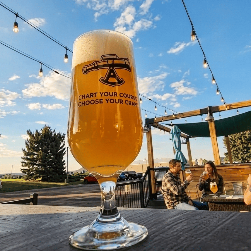 Golden hazy craft beer in an Alidade Brewing glass on the outdoor patio, featuring a foamy head, string lights, and guests enjoying the brewery atmosphere at sunset.