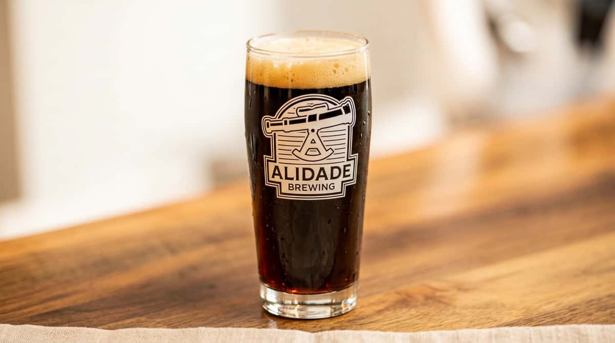 Dark craft beer in an Alidade Brewing pint glass featuring a rich caramel-colored head, served on a wooden bar top and showcasing the brewery's signature stout or porter style.
