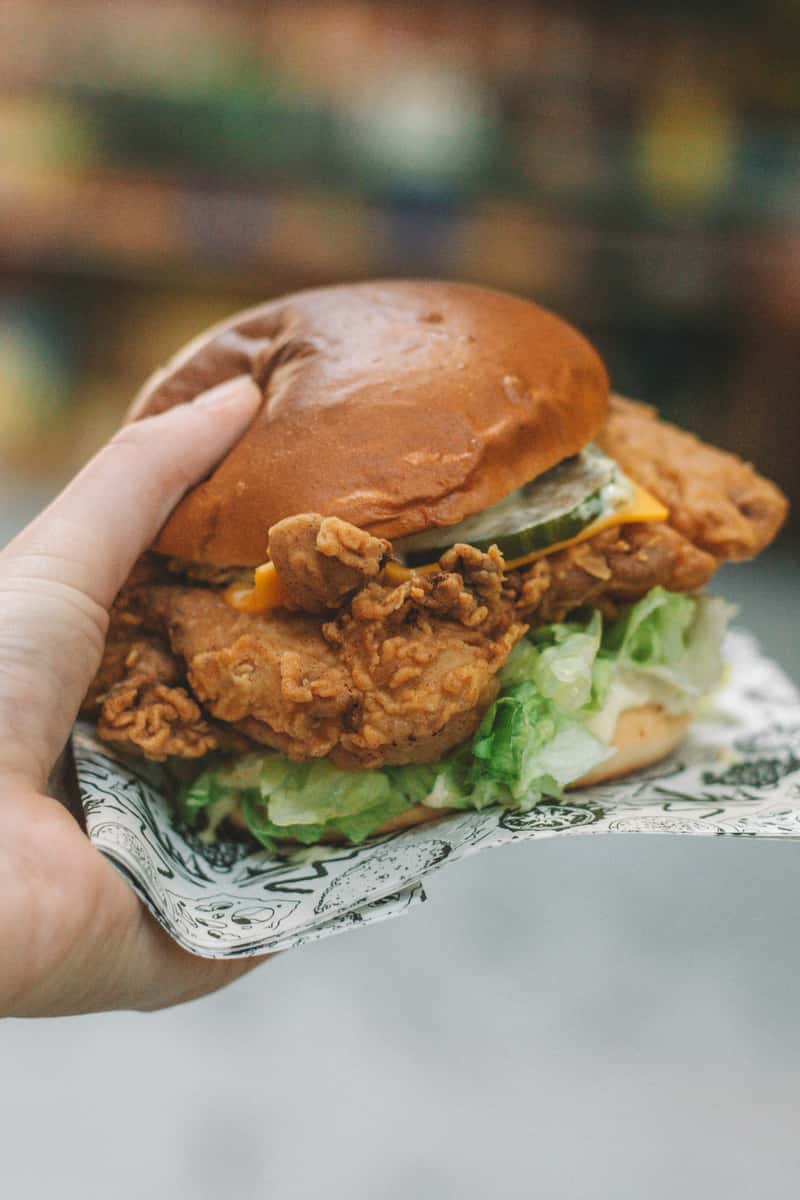 Hand holding a loaded fried chicken sandwich with lettuce, pickles, cheddar cheese, and a soft brioche bun, ideal for gourmet fast food menus.