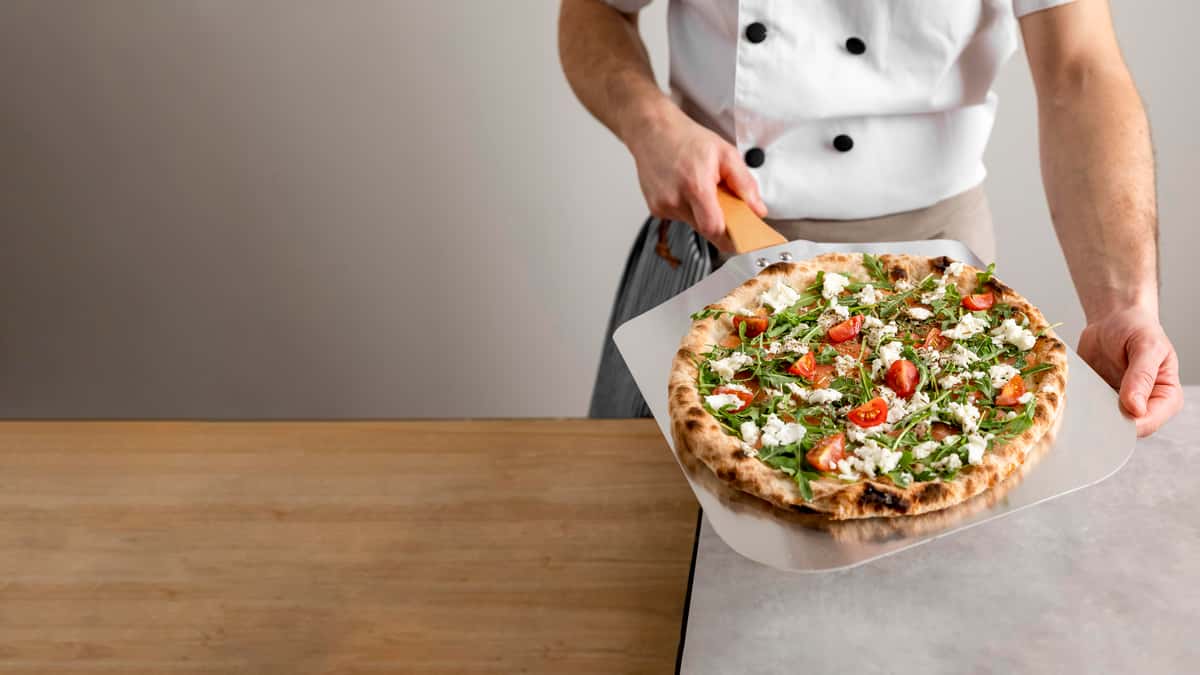 Cheezy Pizza's chef presents a delicious-looking pizza on a peel.