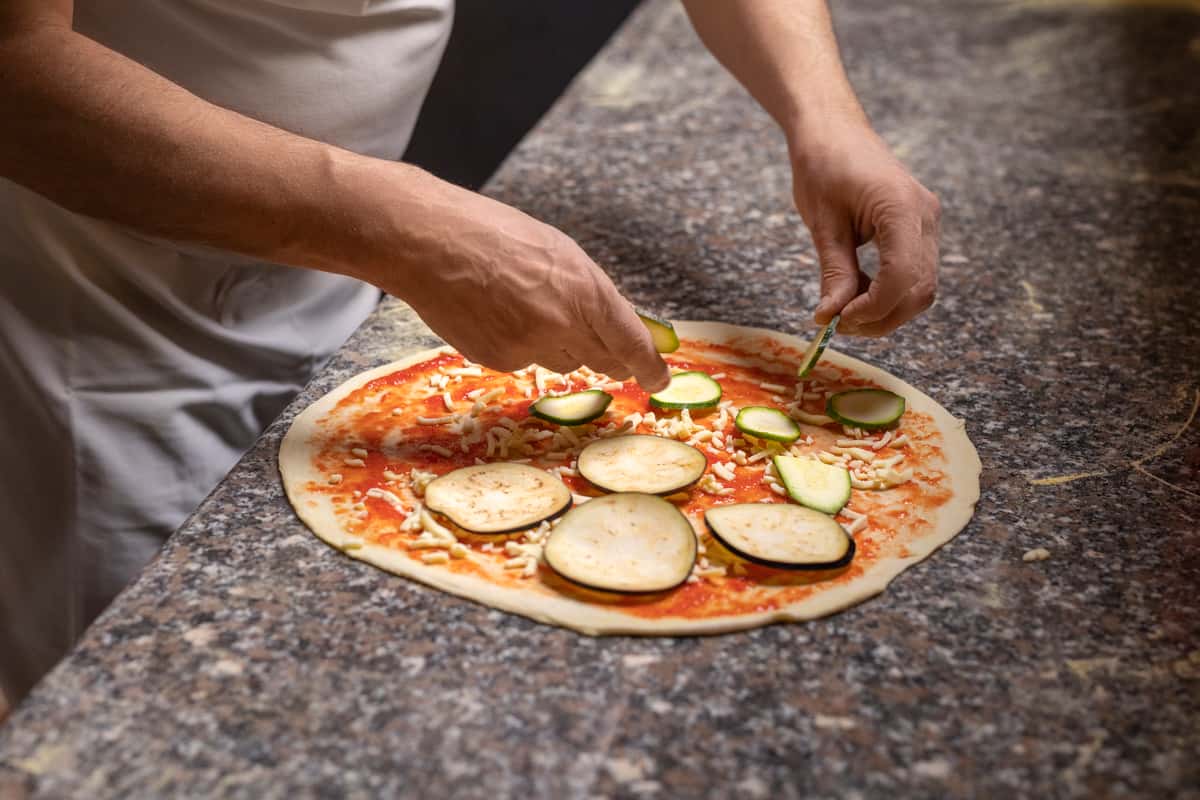 Cheezy Pizza's cheff carefully adds sliced vegetables to a pizza base.