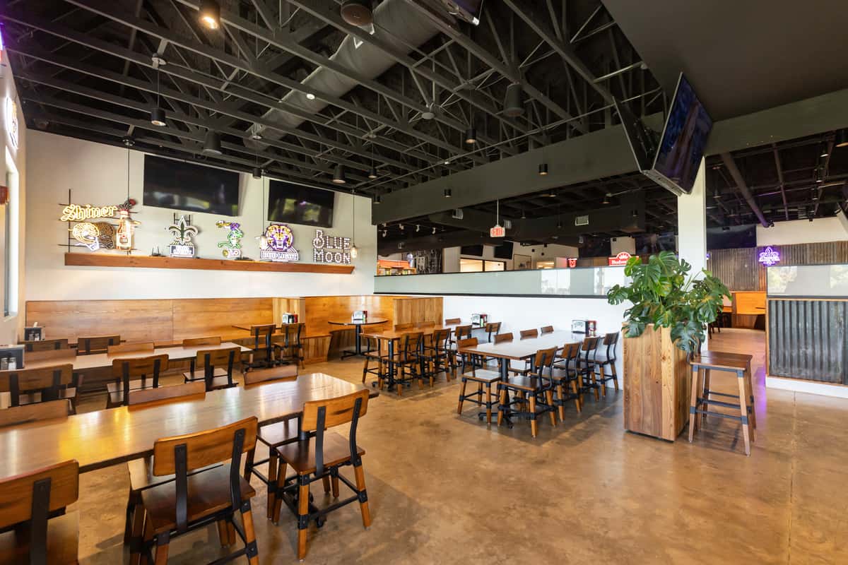 Spacious restaurant interior with wooden tables, neon beer signs, TV screens, and exposed industrial ceiling.