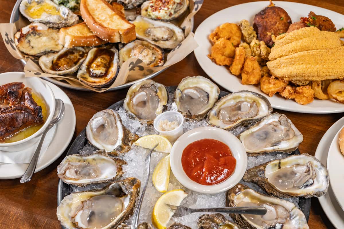 Seafood platter with raw oysters on ice, cocktail sauce, grilled oysters, fried fish, shrimp, and hush puppies on a wooden table.