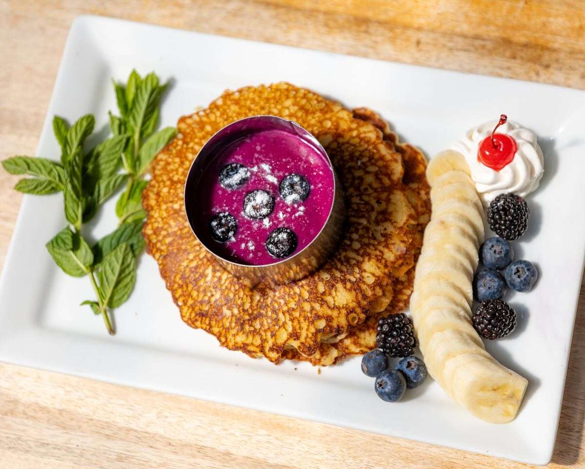 Golden brunch pancakes topped with blueberry sauce, fresh berries, banana slices, whipped cream, and mint garnish