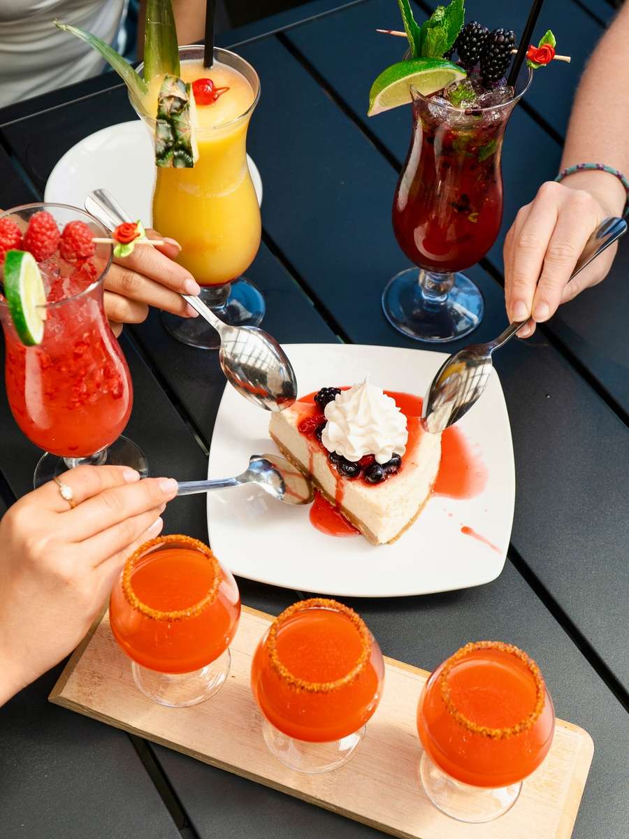 Tropical cocktails, berry mojitos, and spicy margarita shots served with berry cheesecake dessert at a lively outdoor restaurant table