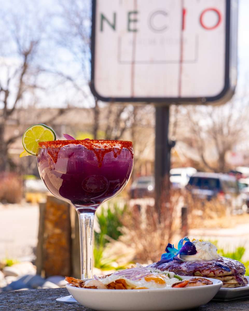 Purple margarita with tajin rim and lime garnish served outdoors at Necio Mexican Kitchen brunch table with pancakes and breakfast plate