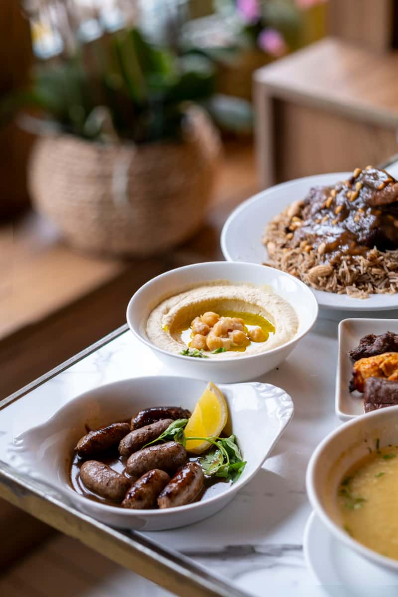 Mediterranean feast with grilled sausages, hummus, lamb shank, and warm soup served on a marble dining table
