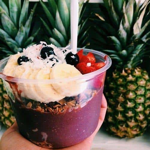 Acai bowl with granola, blueberry, banana, strawberry and shredded coconut against backdrop of whole pineapples.