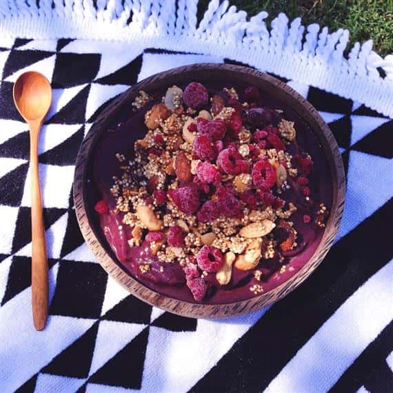 Wooden acai bowl with nuts and raspberries on black and white checkered blanket next to wooden spoon.