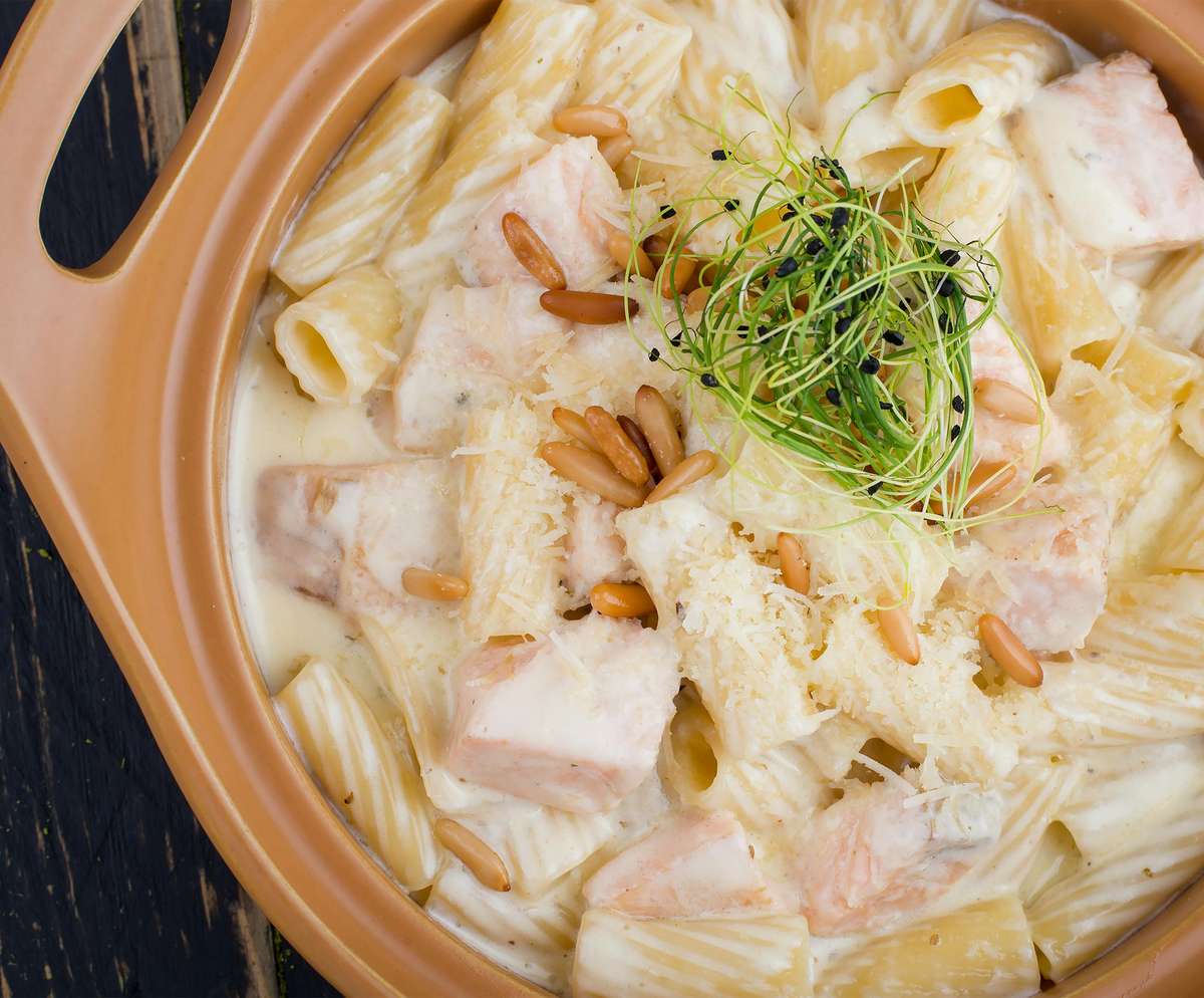 Creamy rigatoni pasta with chunks of salmon, pine nuts, grated cheese, and microgreens in a terracotta serving dish.