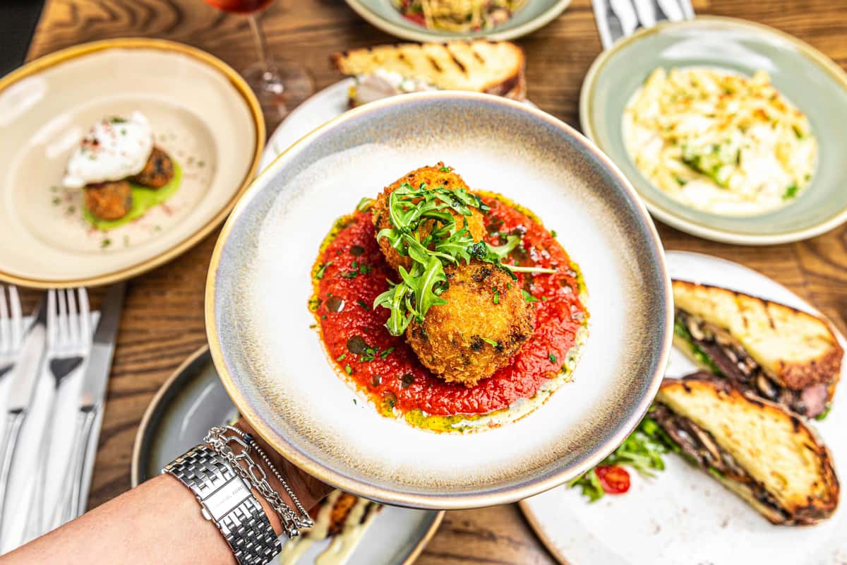Golden arancini balls on rich tomato sauce, garnished with arugula and herbs, held above a table of assorted Italian dishes.