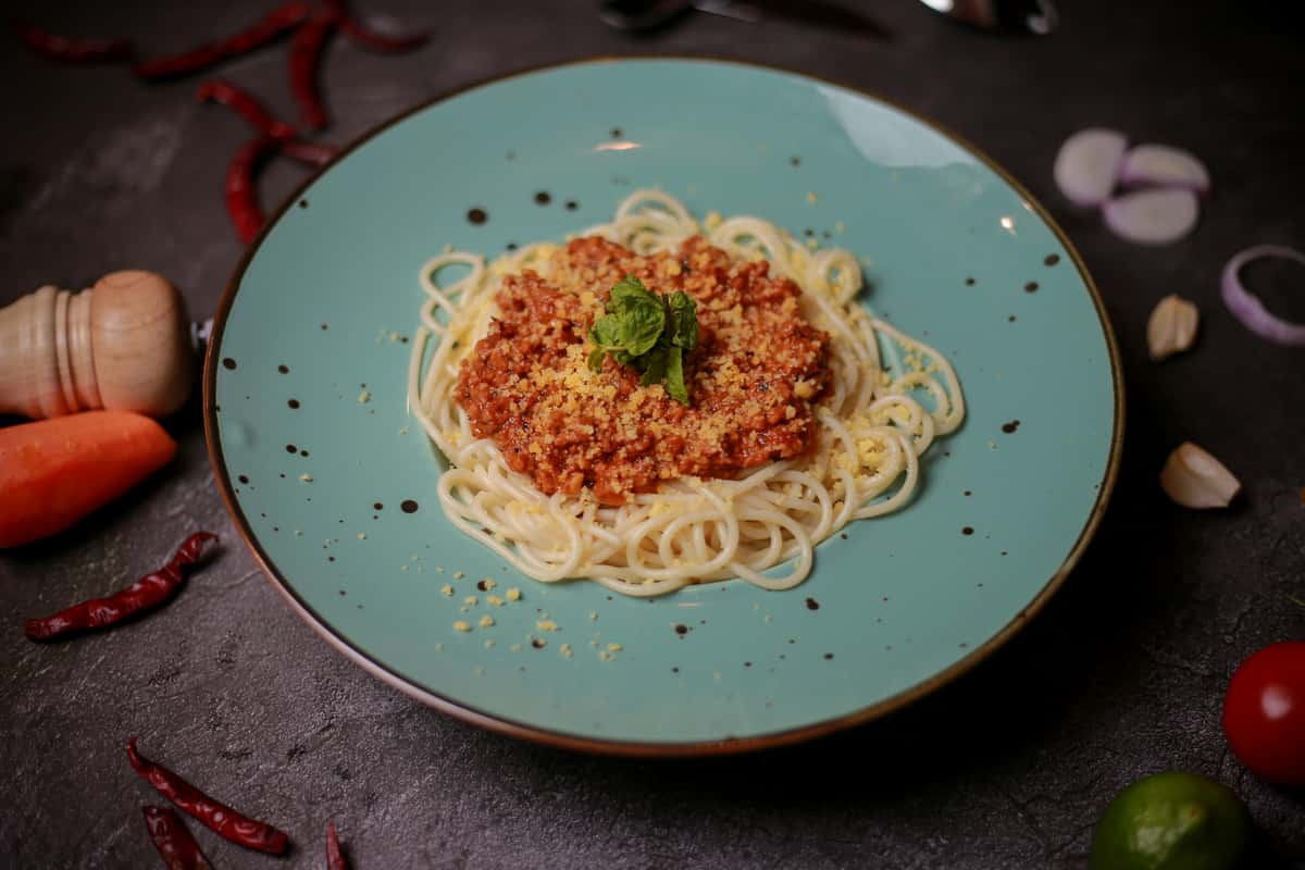 Spaghetti topped with bolognese sauce, grated cheese, and basil, served on a turquoise plate with scattered ingredients.