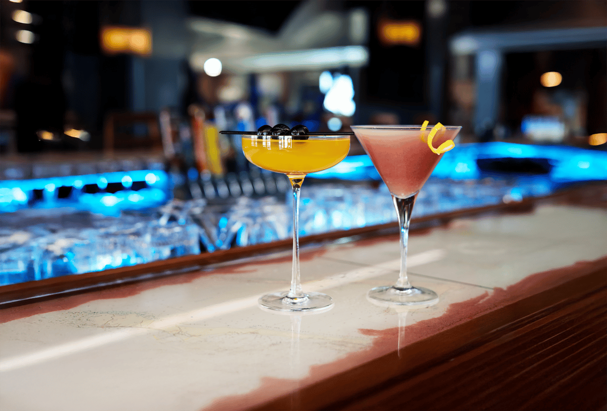 Two elegant cocktails sit on a polished bar. A vibrant yellow drink garnished with blueberries and a pink martini with a lemon twist