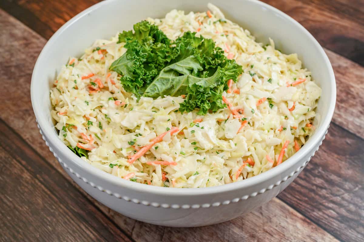 Creamy coleslaw with shredded cabbage and carrots, topped with leafy green garnish in a white bowl.