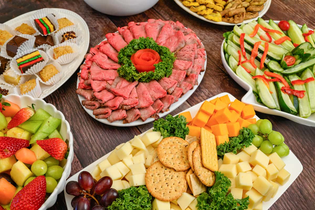 Buffet table with roast beef, cheese cubes, fresh fruit, cucumber salad, and assorted bite-sized desserts.