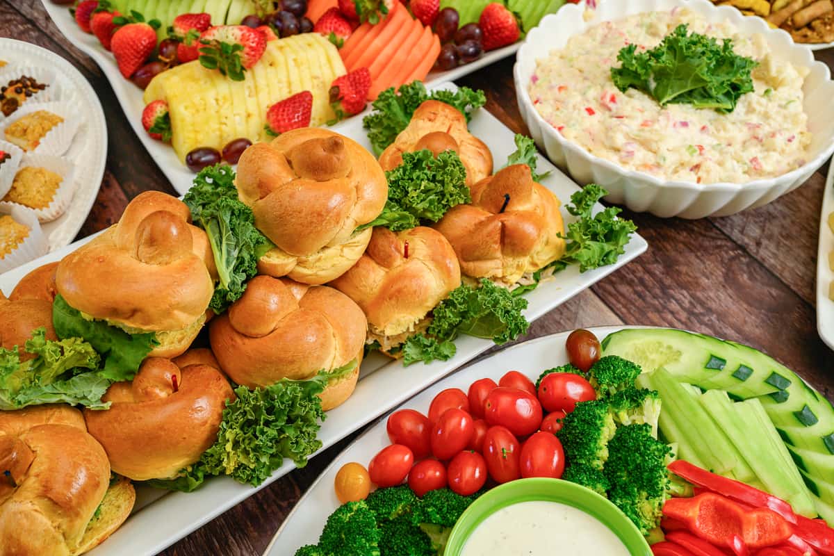 Party platter with mini sandwiches, fresh vegetable tray with dip, fruit slices, and a bowl of creamy potato salad on a wooden table.